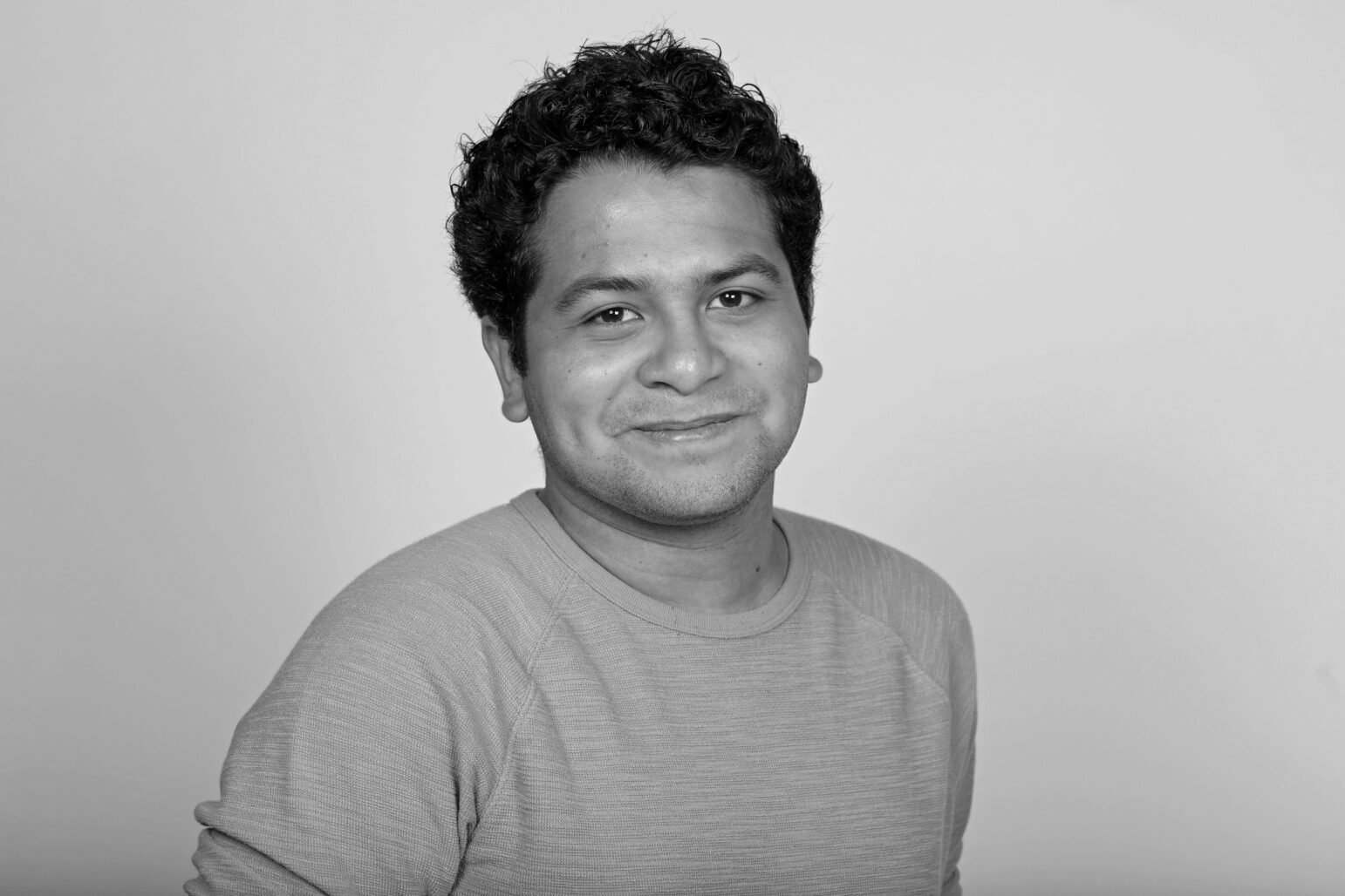 A black-and-white portrait of a man with short, curly hair, wearing a light sweater and smiling softly against a plain background.