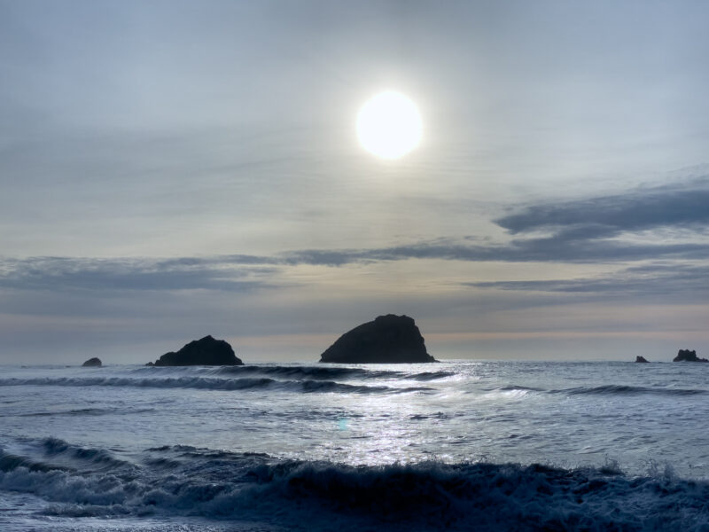 The sun shines over the Pacific Ocean near Eureka, California, casting a reflective glow across gentle waves and silhouetting rocky outcroppings against a soft, overcast sky.