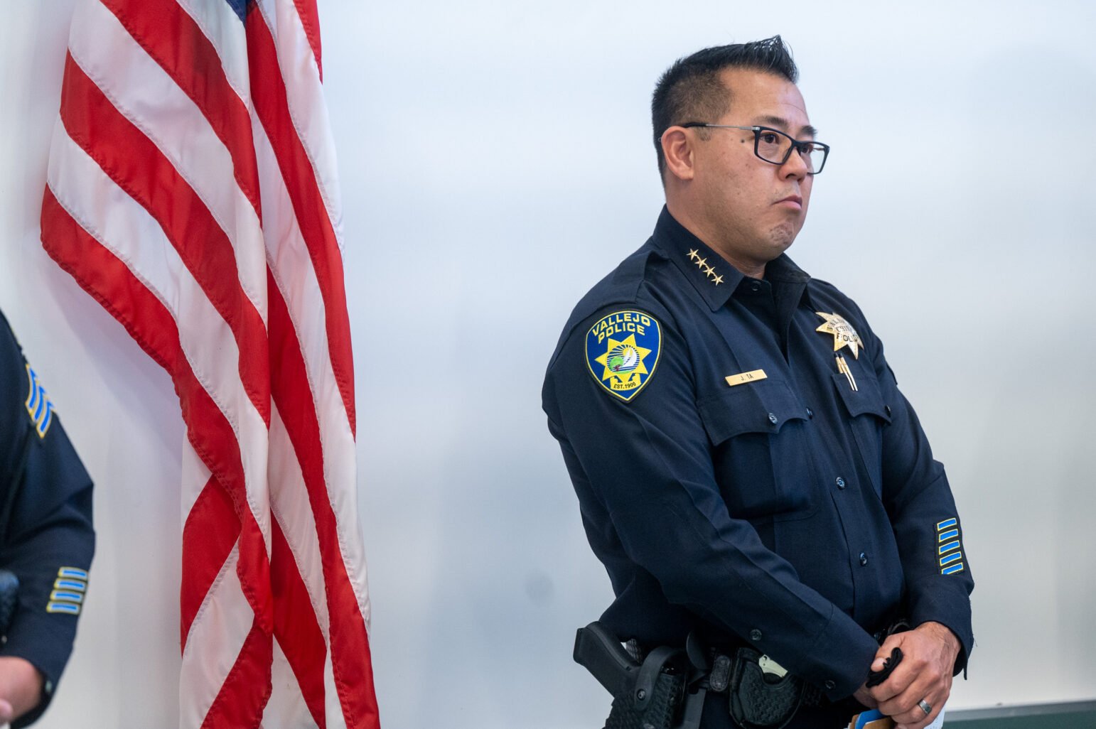 A Vallejo police officer in full uniform, identified by the badge on his shirt as Jason Ta, stands next to an American flag. His expression is serious, and he is positioned against a plain background, with the department's patch visible on his sleeve.