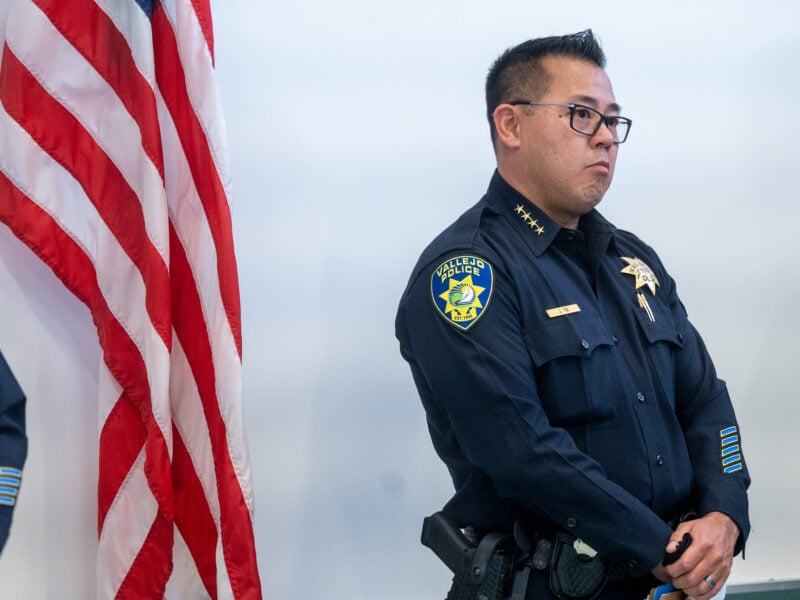 A Vallejo police officer in full uniform, identified by the badge on his shirt as Jason Ta, stands next to an American flag. His expression is serious, and he is positioned against a plain background, with the department's patch visible on his sleeve.