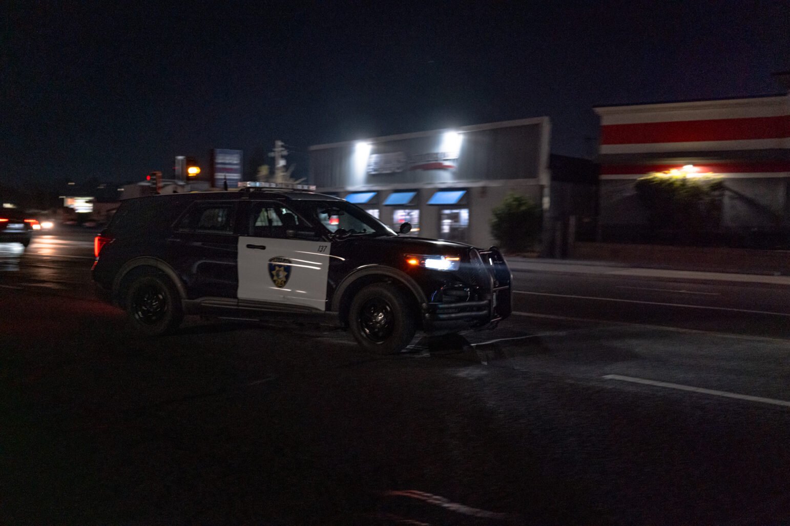 A Vallejo Police Department SUV is captured driving through a dimly lit street at night, with storefronts illuminated in the background.