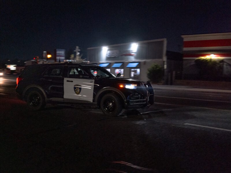 A Vallejo Police Department SUV is captured driving through a dimly lit street at night, with storefronts illuminated in the background.