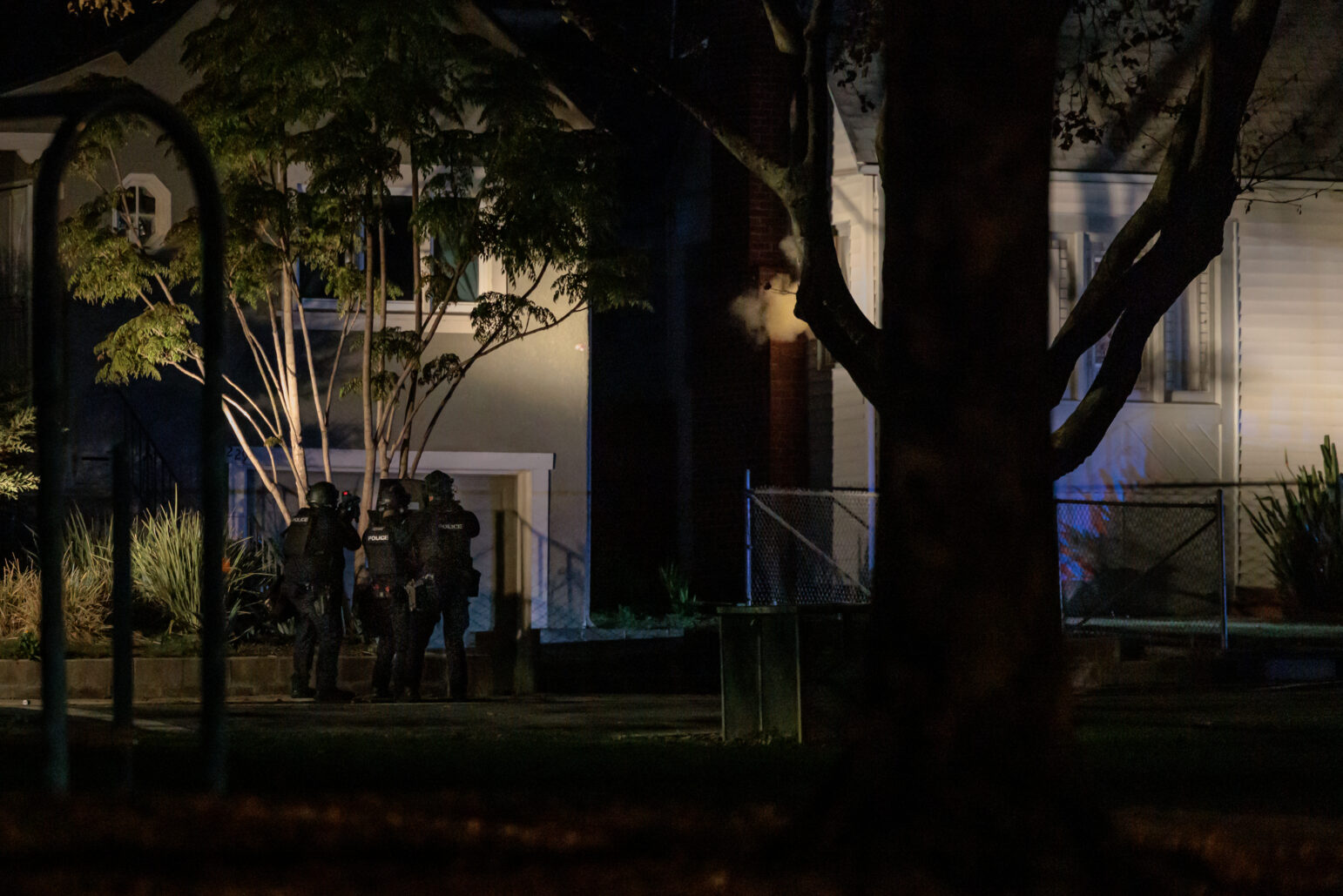 Three SWAT officers, wearing tactical gear, approach a house at night. The house is partially obscured by trees, and a faint cloud of gas or smoke is visible near the structure. The scene is illuminated by artificial lighting, casting dramatic shadows on the house and surrounding foliage. A metal fence and neighboring homes are visible in the background.