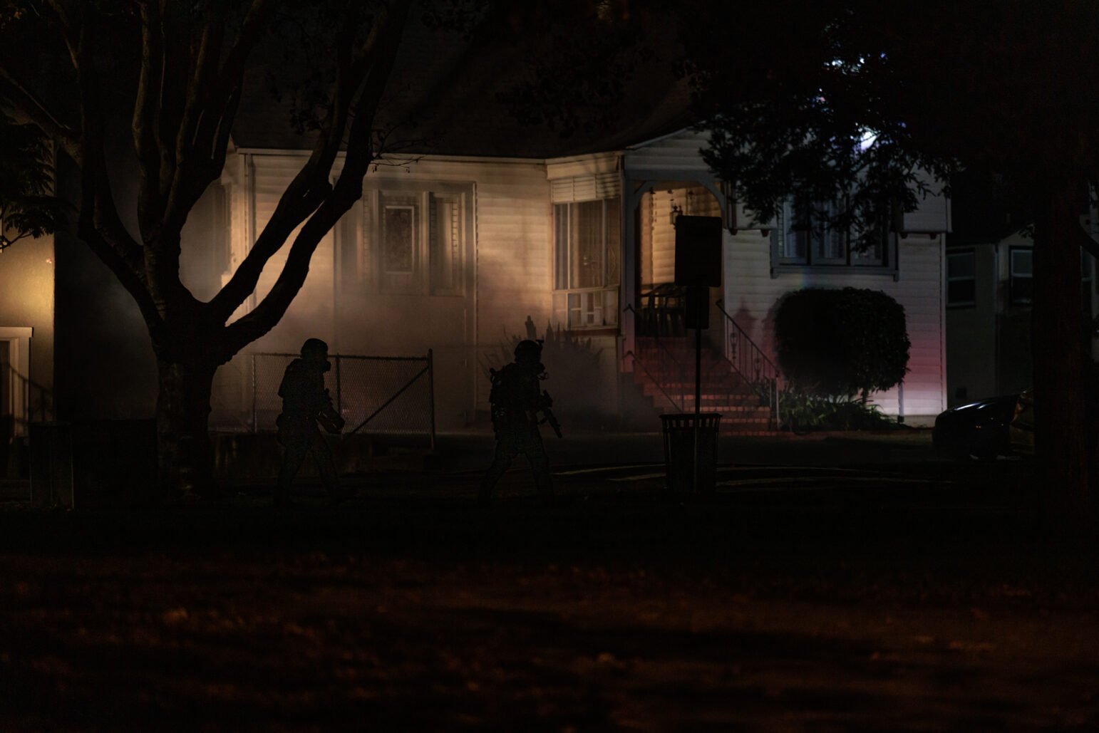 Silhouetted SWAT officers move cautiously across a yard in front of a house at night, partially obscured by a dense cloud of gas. The scene is illuminated by ambient streetlights and faint reflections from nearby emergency vehicles, casting long shadows on the house and surrounding trees. The home's front porch and staircase are faintly visible, framed by foliage and a chain-link fence.