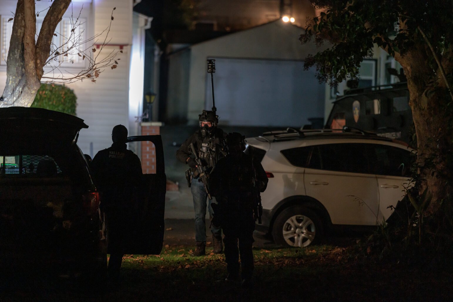 Silhouetted police officers stand near a vehicle in a residential driveway at night, one officer partially visible in tactical gear, holding a firearm. A white SUV and a specialized armored police vehicle are parked nearby, illuminated by ambient light from the house and surrounding area. A bare tree and fallen leaves frame the scene, adding depth.