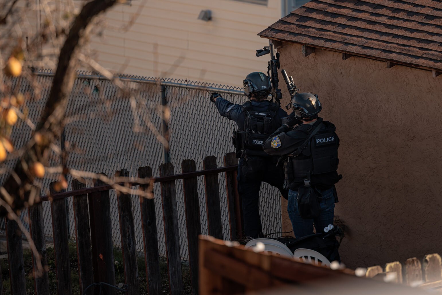 Two police officers in tactical gear, identified as members of a SWAT team, assess the area during an operation. One officer gestures forward while the other holds a rifle, both positioned near a chain-link fence and a small structure, partially obscured by a tree branch in the foreground.