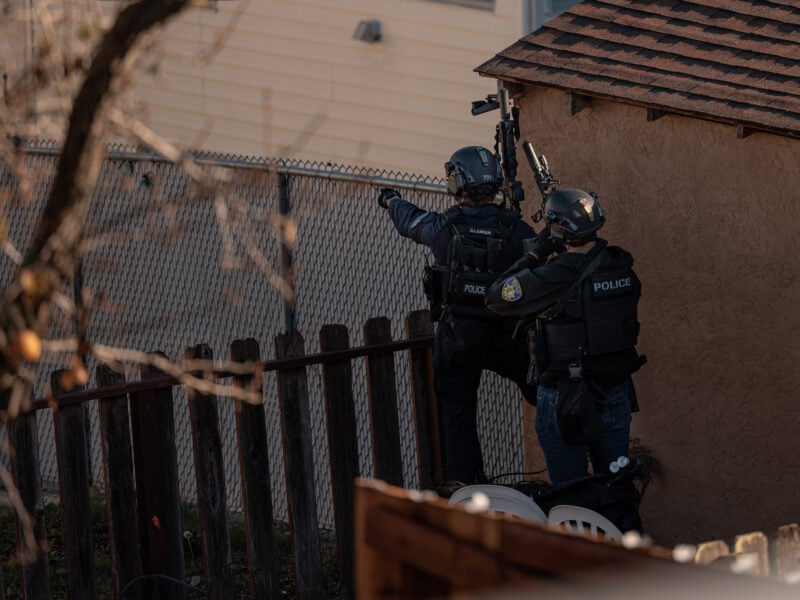 Two police officers in tactical gear, identified as members of a SWAT team, assess the area during an operation. One officer gestures forward while the other holds a rifle, both positioned near a chain-link fence and a small structure, partially obscured by a tree branch in the foreground.