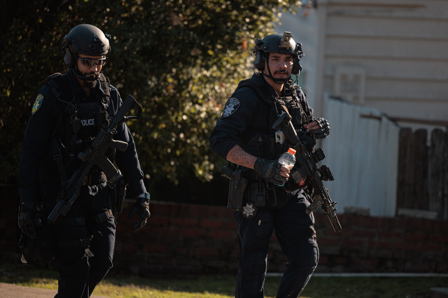 Two SWAT officers walk side by side during an operation. Both are dressed in tactical gear, carrying rifles and wearing helmets equipped with communication devices. One officer holds a Gatorade bottle, and sunlight filters through trees in the background, partially illuminating their path. A residential fence and brick wall are visible in the background.