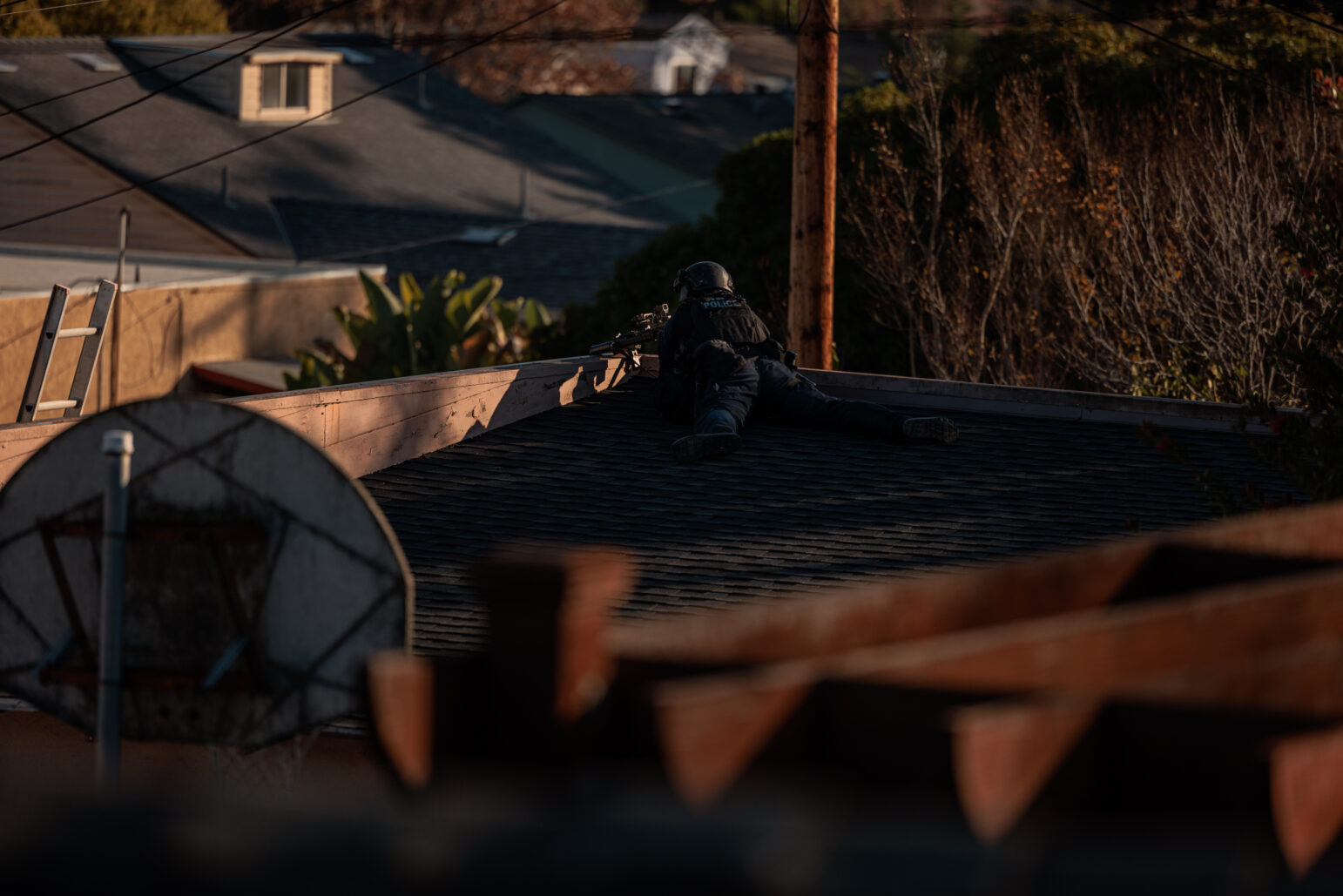 A SWAT officer lies prone on the roof of a house, aiming a rifle while dressed in tactical gear. The setting is a residential neighborhood with rooftops, power lines, and vegetation visible in the background. A residential satellite dish and a wooden structure are partially in the foreground, framing the scene. The late afternoon sunlight casts long shadows, highlighting the officer's focused position.