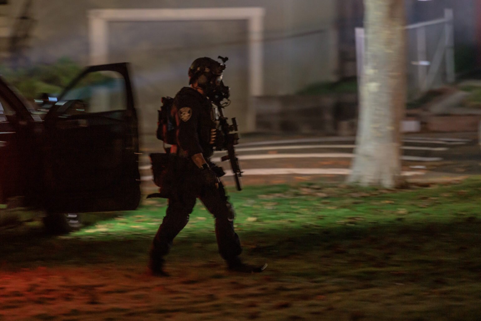 A SWAT officer in tactical gear walks across a grassy area at night, carrying a rifle. The scene is dimly lit with a mix of artificial light sources, creating a sense of urgency. An open vehicle door is visible in the background, along with blurred elements of a residential setting, including a tree and faint outlines of a house.