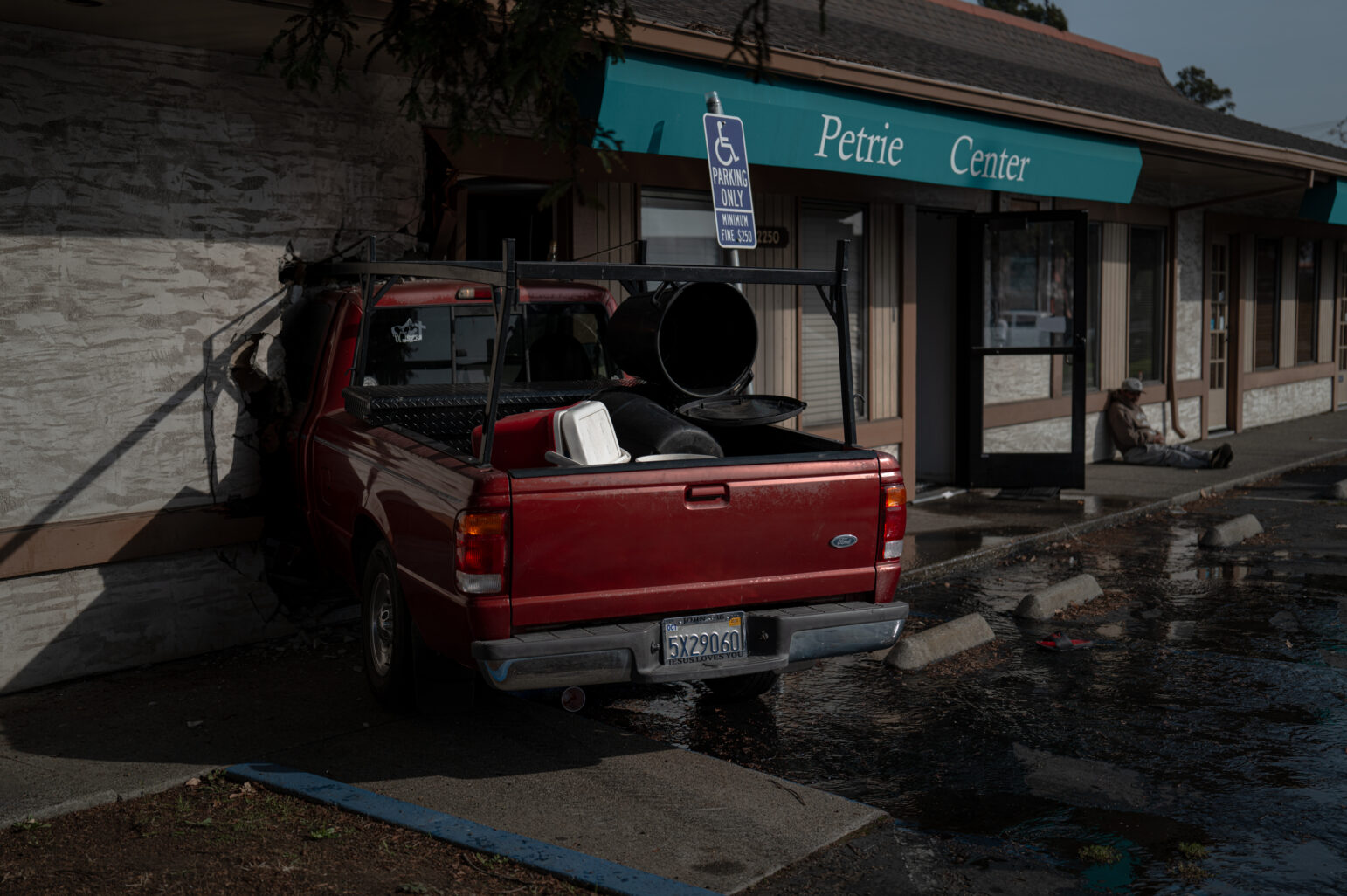 The red pickup truck remains embedded in the Petrie Center wall, with debris scattered around the parking lot. A man sits near the building’s entrance.