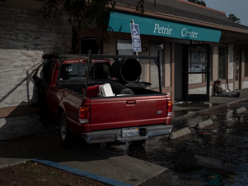 The red pickup truck remains embedded in the Petrie Center wall, with debris scattered around the parking lot. A man sits near the building’s entrance.