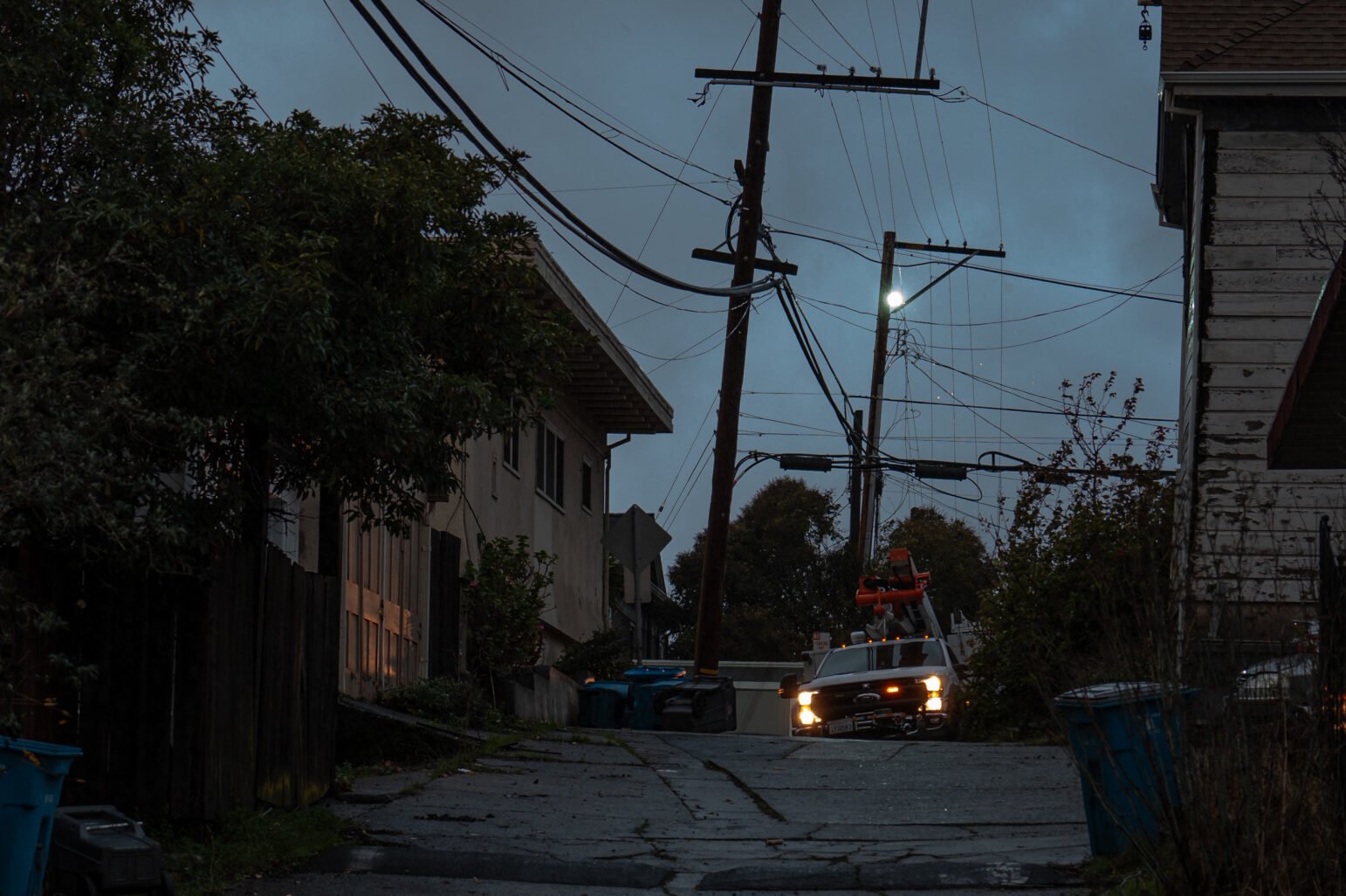 A dimly lit alley with overhead power lines, some leaning precariously, as a utility truck with headlights on is parked near the base of the lines. Nearby, blue recycling bins and vegetation line the pathway, and a faint glow from electrical arcing illuminates the evening sky.