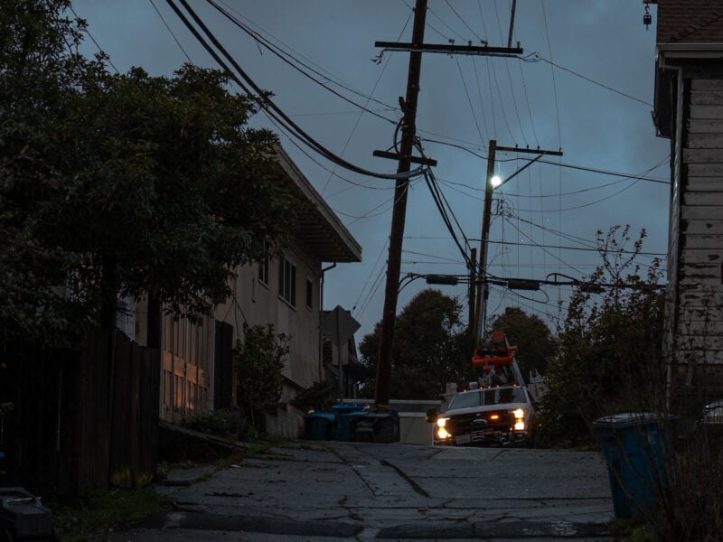 A dimly lit alley with overhead power lines, some leaning precariously, as a utility truck with headlights on is parked near the base of the lines. Nearby, blue recycling bins and vegetation line the pathway, and a faint glow from electrical arcing illuminates the evening sky.