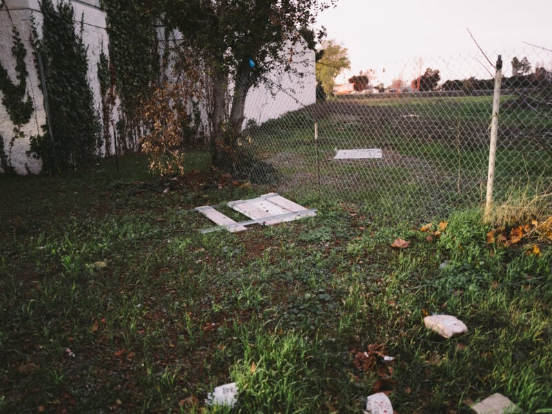 A chain-link fence with a broken section lying on the grass, surrounded by scattered debris and overgrown vegetation. Ivy climbs a nearby building wall, and an open, grassy lot extends into the distance under the soft light of dusk.