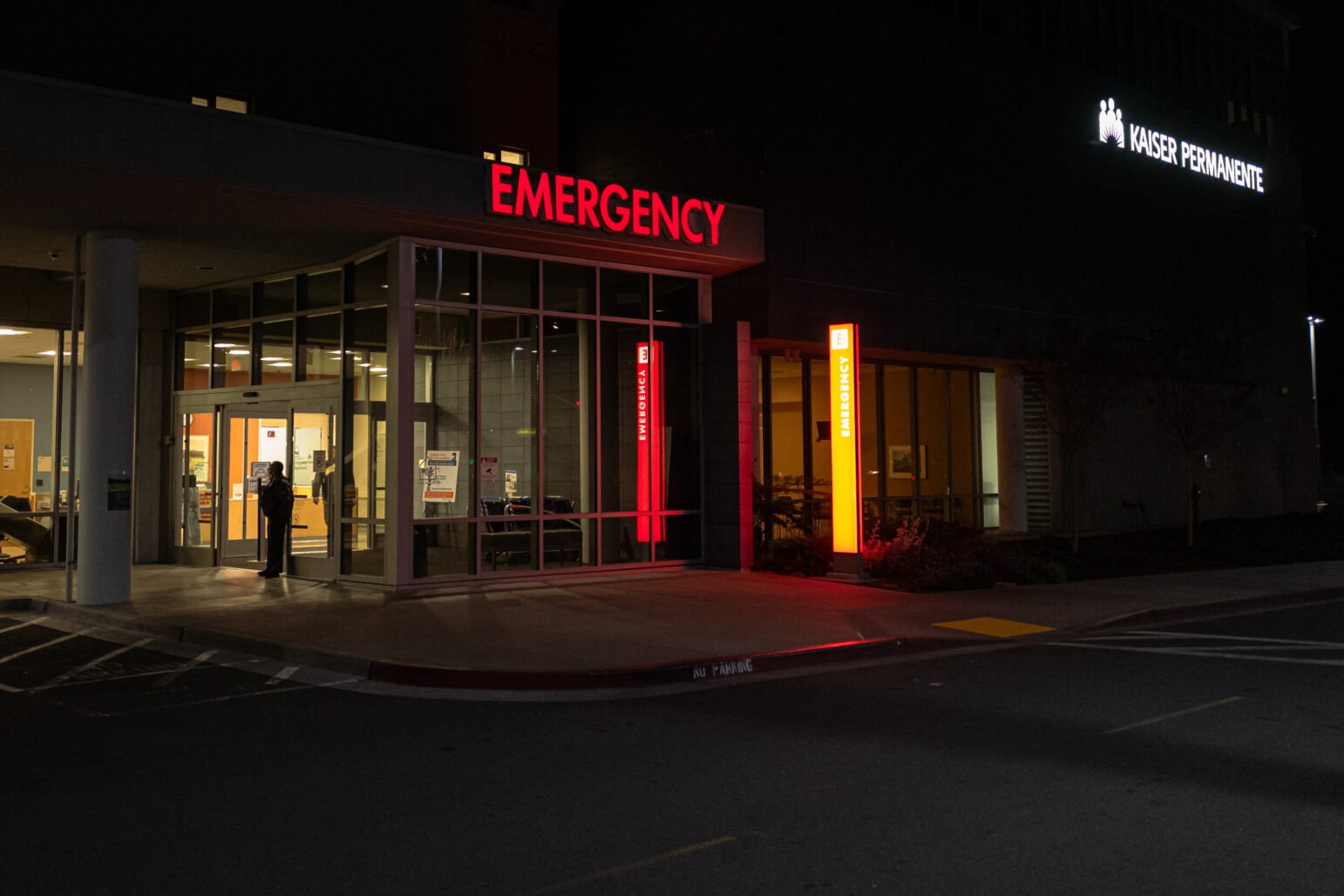 The entrance to the emergency room at Kaiser Permanente in Vallejo, illuminated by bright red "EMERGENCY" signage against the dark night. A person is visible standing near the glass doors, adding a sense of quiet urgency.