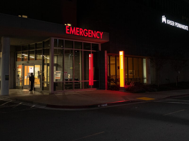 The entrance to the emergency room at Kaiser Permanente in Vallejo, illuminated by bright red "EMERGENCY" signage against the dark night. A person is visible standing near the glass doors, adding a sense of quiet urgency.