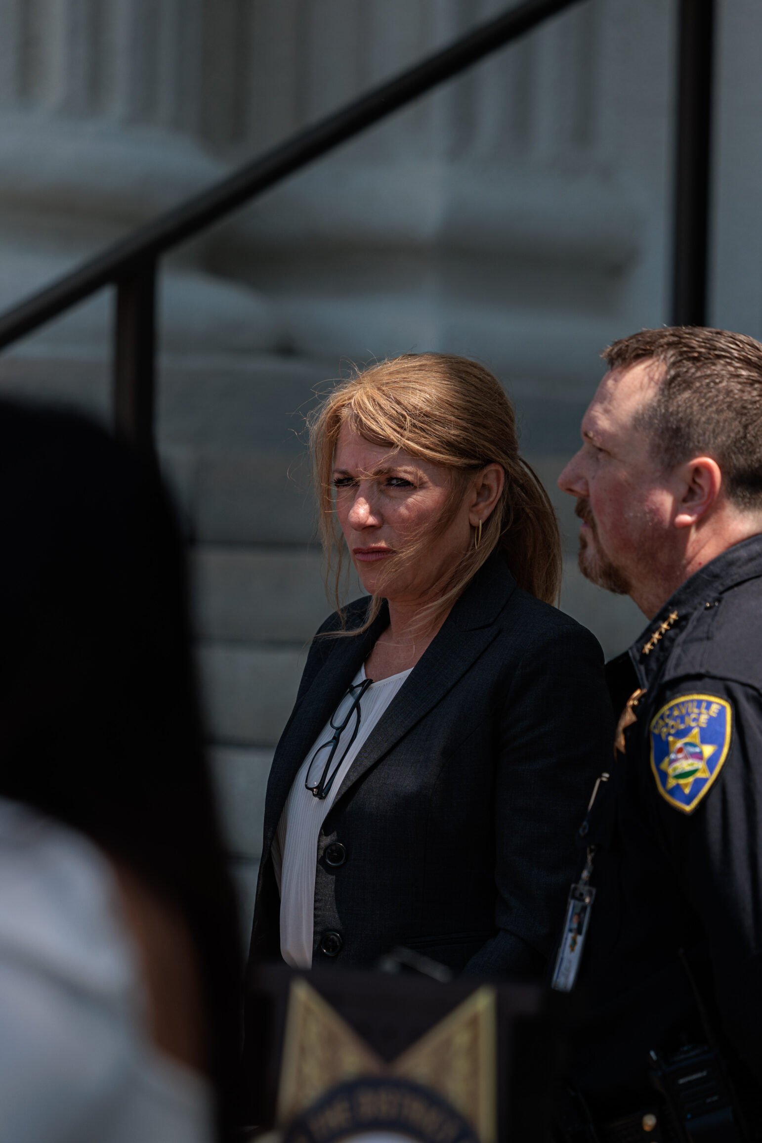 A woman with blond hair, wearing a dark blazer and a white blouse, stands on the steps of a building next to a uniformed police officer. She appears focused and a pair of glasses hangs from her collar. The officer, in a dark police uniform, is partially turned toward her. The building’s columns and handrail are visible in the background.