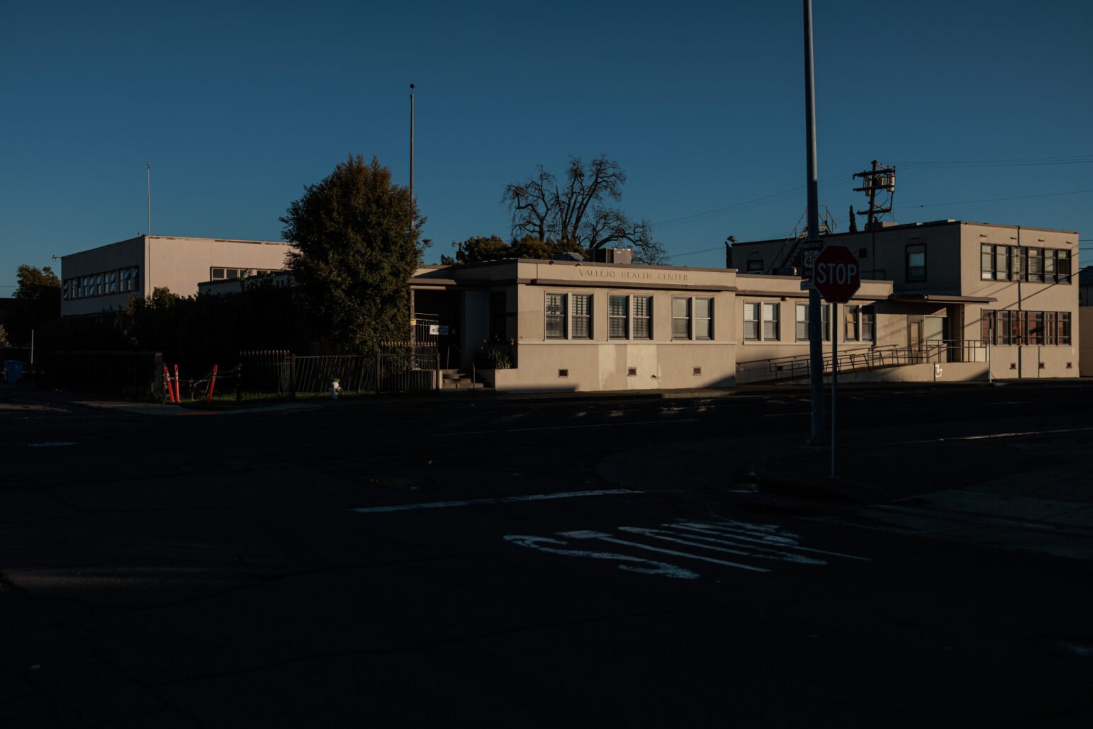 A distant shot of the Vallejo Health Center in the early evening, with the building partially lit by the setting sun, surrounded by shadows and empty streets.