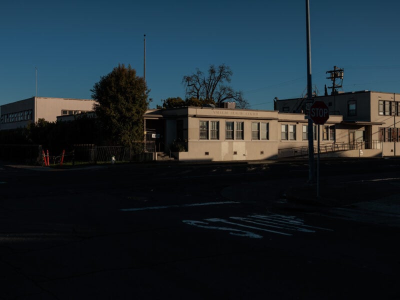 A distant shot of the Vallejo Health Center in the early evening, with the building partially lit by the setting sun, surrounded by shadows and empty streets.