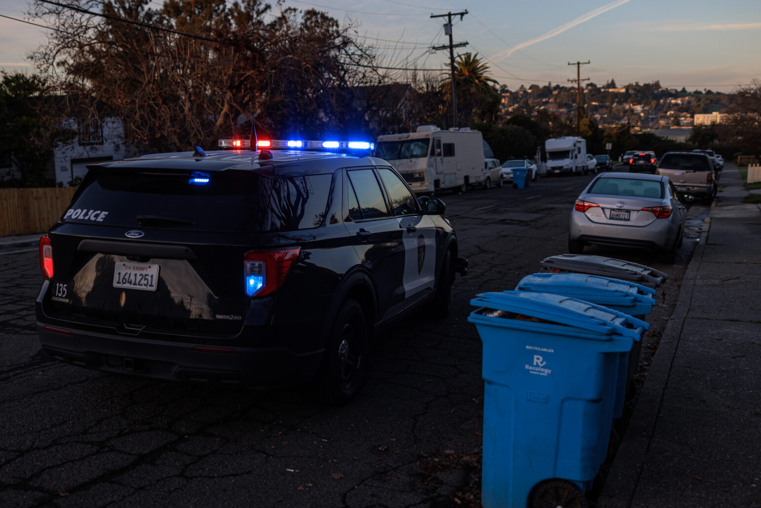 A police vehicle, illuminated by red and blue lights, parked on a residential street lined with recycling bins in morning light.