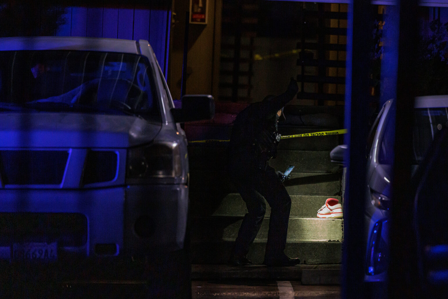 A police investigator examines the scene of a shooting at night, illuminated by blue emergency lights, with a single red-and-white sneaker resting on the steps in the background, partially blocked by parked vehicles.