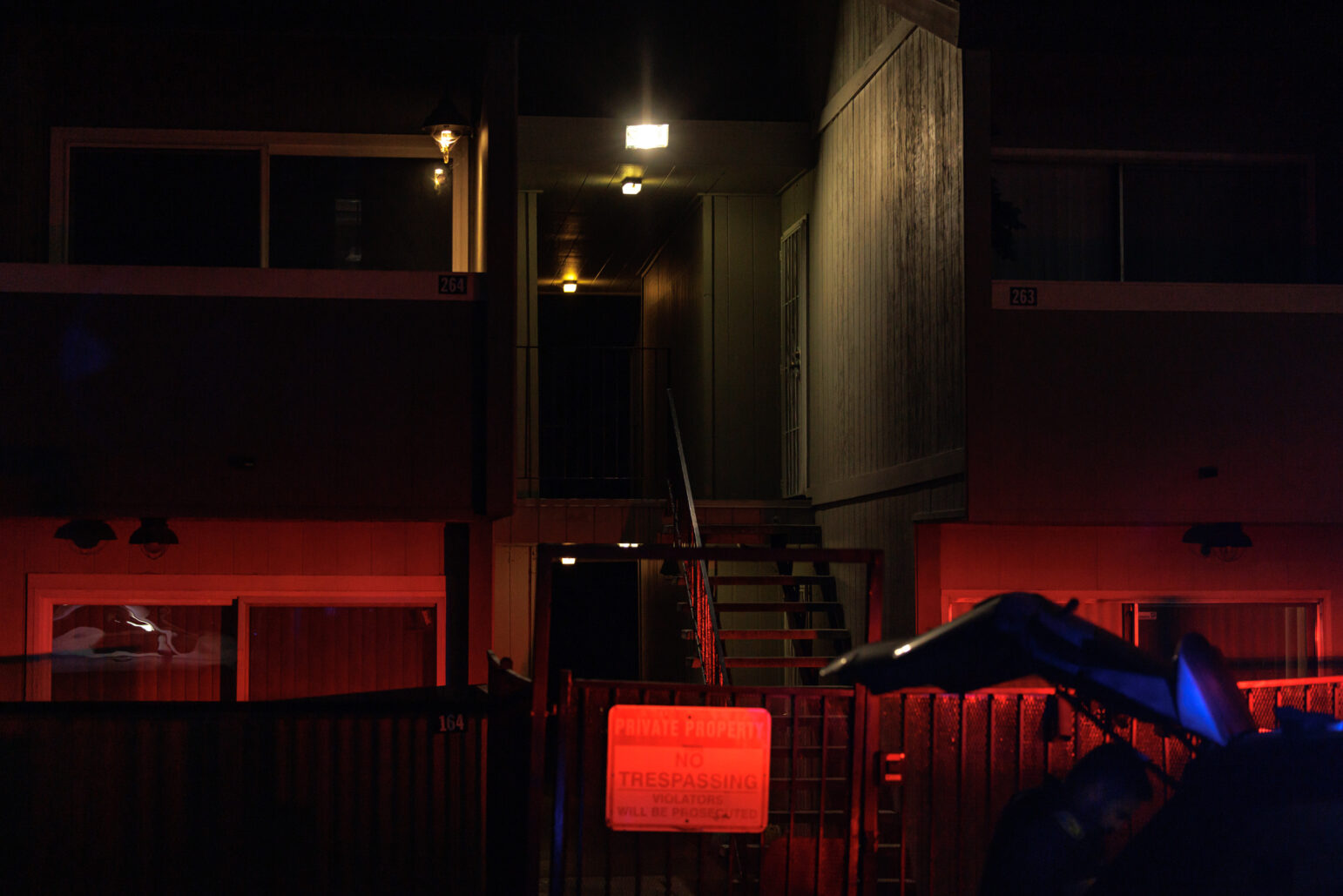 A gated apartment building at night, with a red "No Trespassing" sign visible under the glare of police vehicle lights.