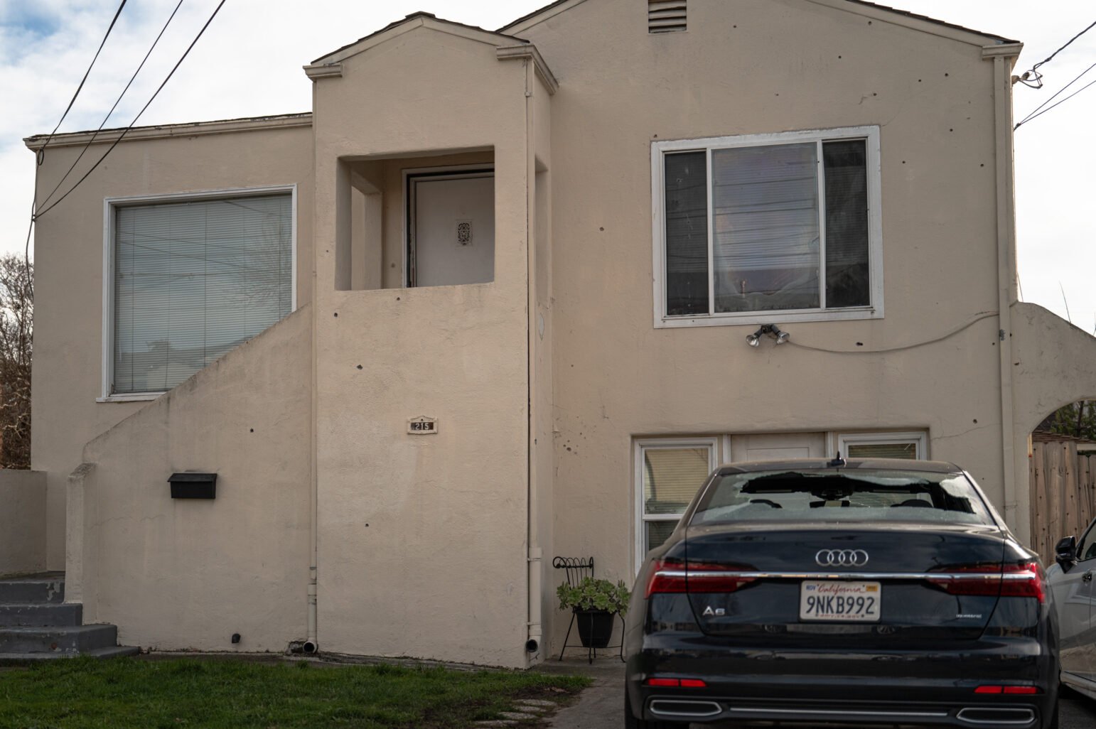 A shattered vehicle windshield and bullet-marked façade of a beige house on a quiet street in daylight.