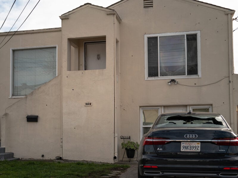 A shattered vehicle windshield and bullet-marked façade of a beige house on a quiet street in daylight.