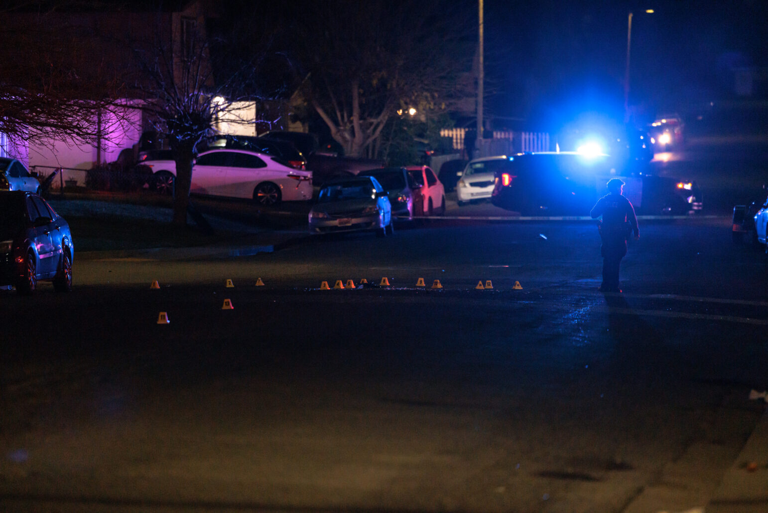 A police officer stands in the distance on a dimly lit street, illuminated by police vehicle lights. Yellow evidence markers are spread across the road, with a police SUV and residential houses in the background.