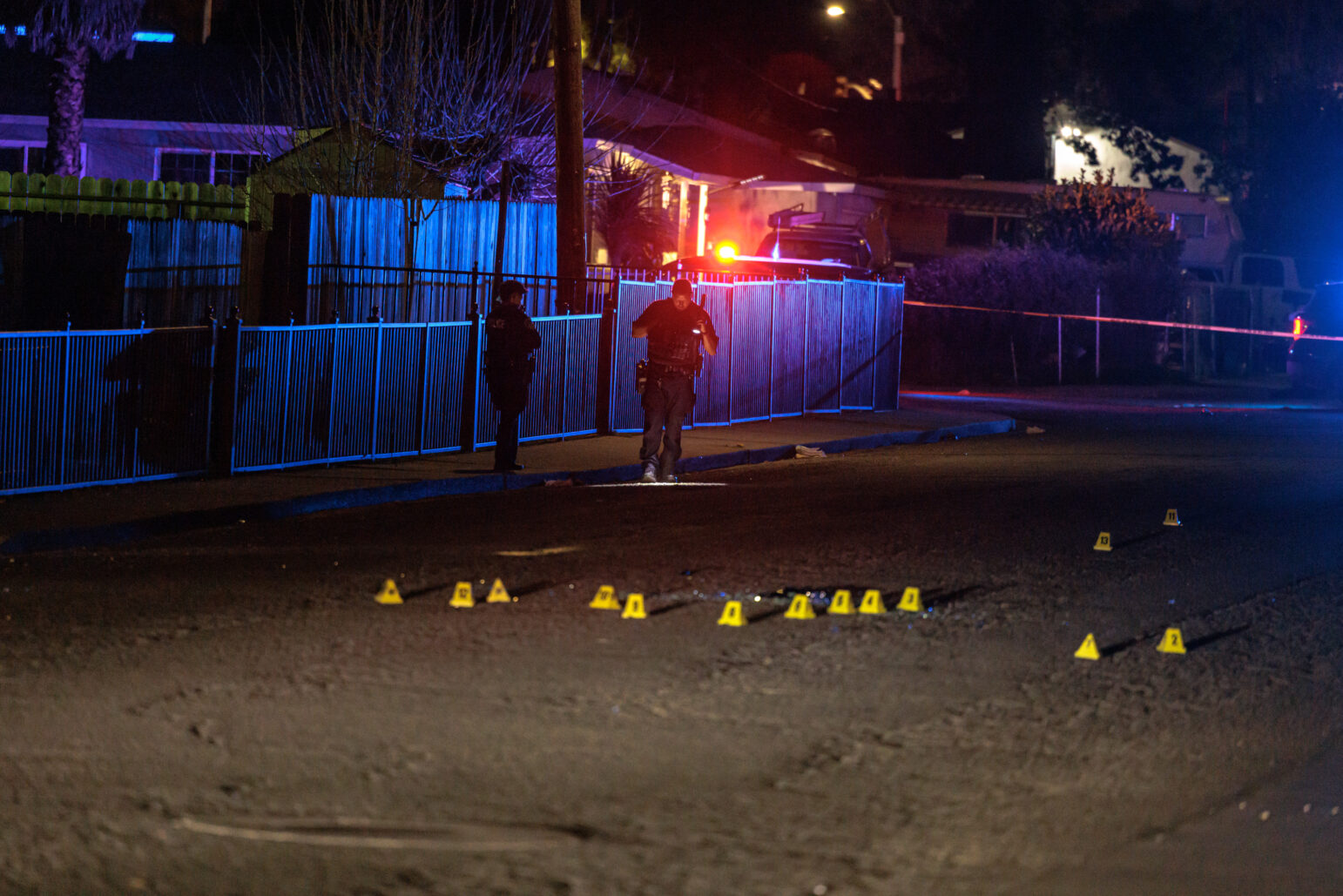 Two police officers walk along a sidewalk beside a blue-lit fence. Yellow evidence markers and scattered debris are visible on the dark street, cordoned off with police tape.