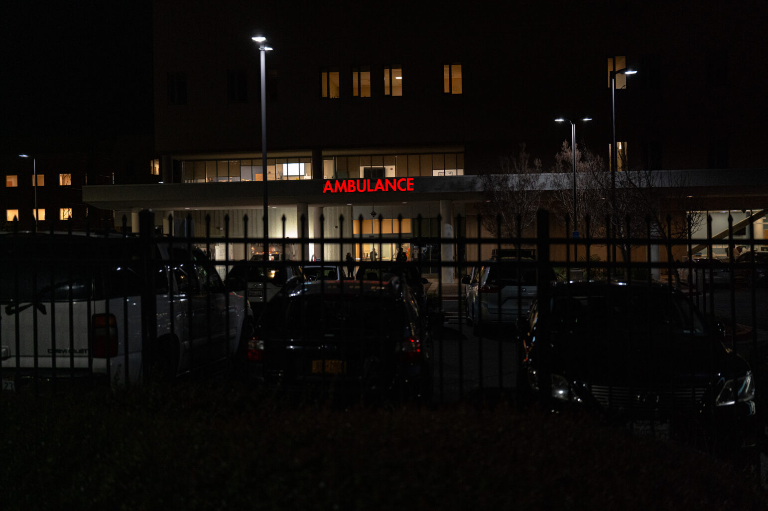 The entrance to a hospital emergency area at night, with the word "AMBULANCE" illuminated in red letters above the entrance. Several parked cars are visible in front of the building, separated by a black metal fence.