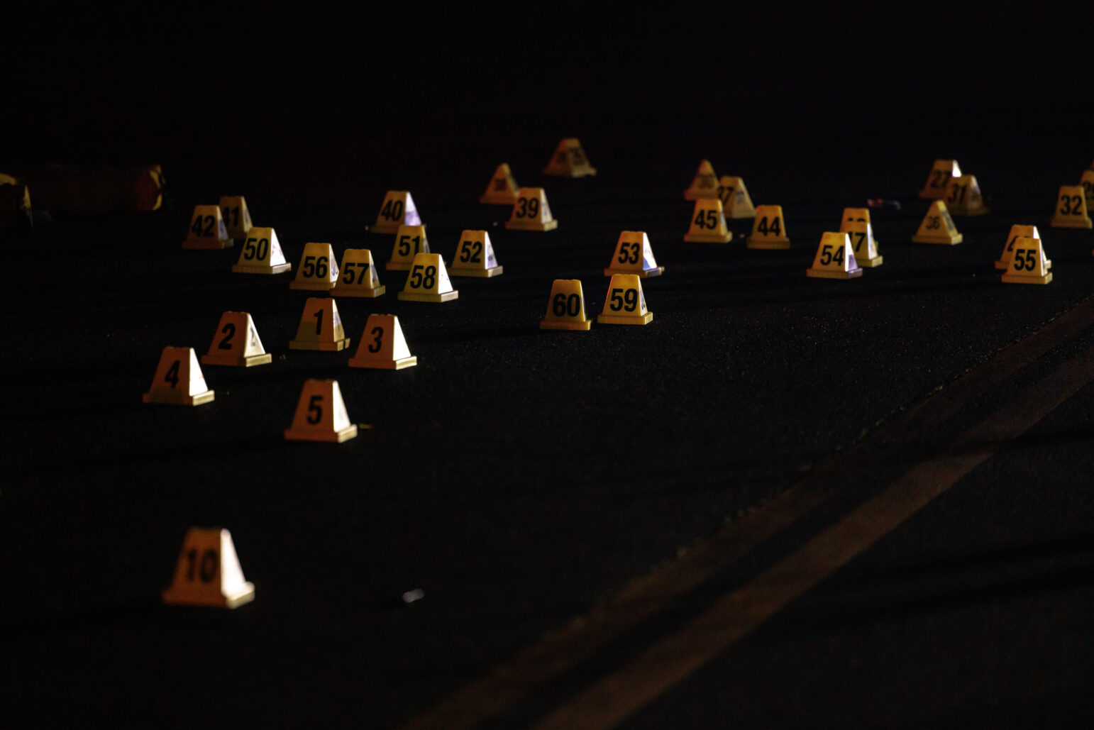 A close-up view of many yellow evidence markers numbered 1 through 60 scattered on the dark pavement, illuminated by artificial light.