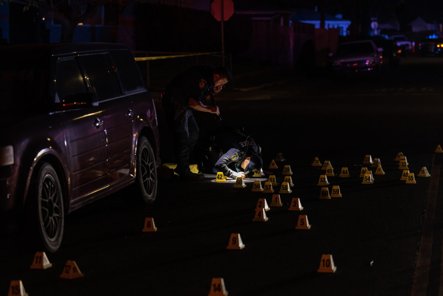 Two police officers examine the ground at night next to a maroon SUV. One officer kneels with a flashlight near numerous yellow evidence markers numbered in the 40s and 50s, while the other stands nearby.