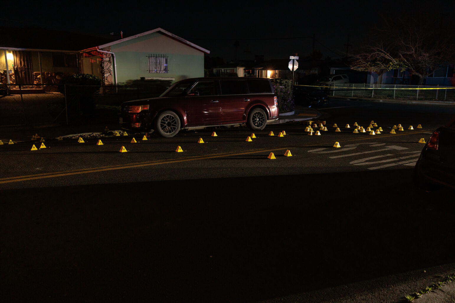 A dark street scene with a maroon SUV parked near the curb. Dozens of yellow evidence markers are scattered across the road, indicating a crime scene. A house with a lit porch is visible in the background.