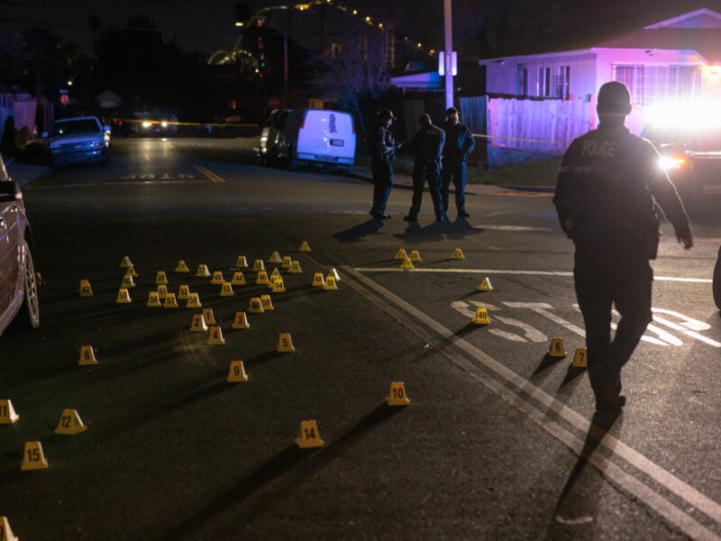 A police officer walks past dozens of yellow evidence markers on a dark street. Three officers stand near a police vehicle in the distance, with flashing lights casting a glow on nearby houses.