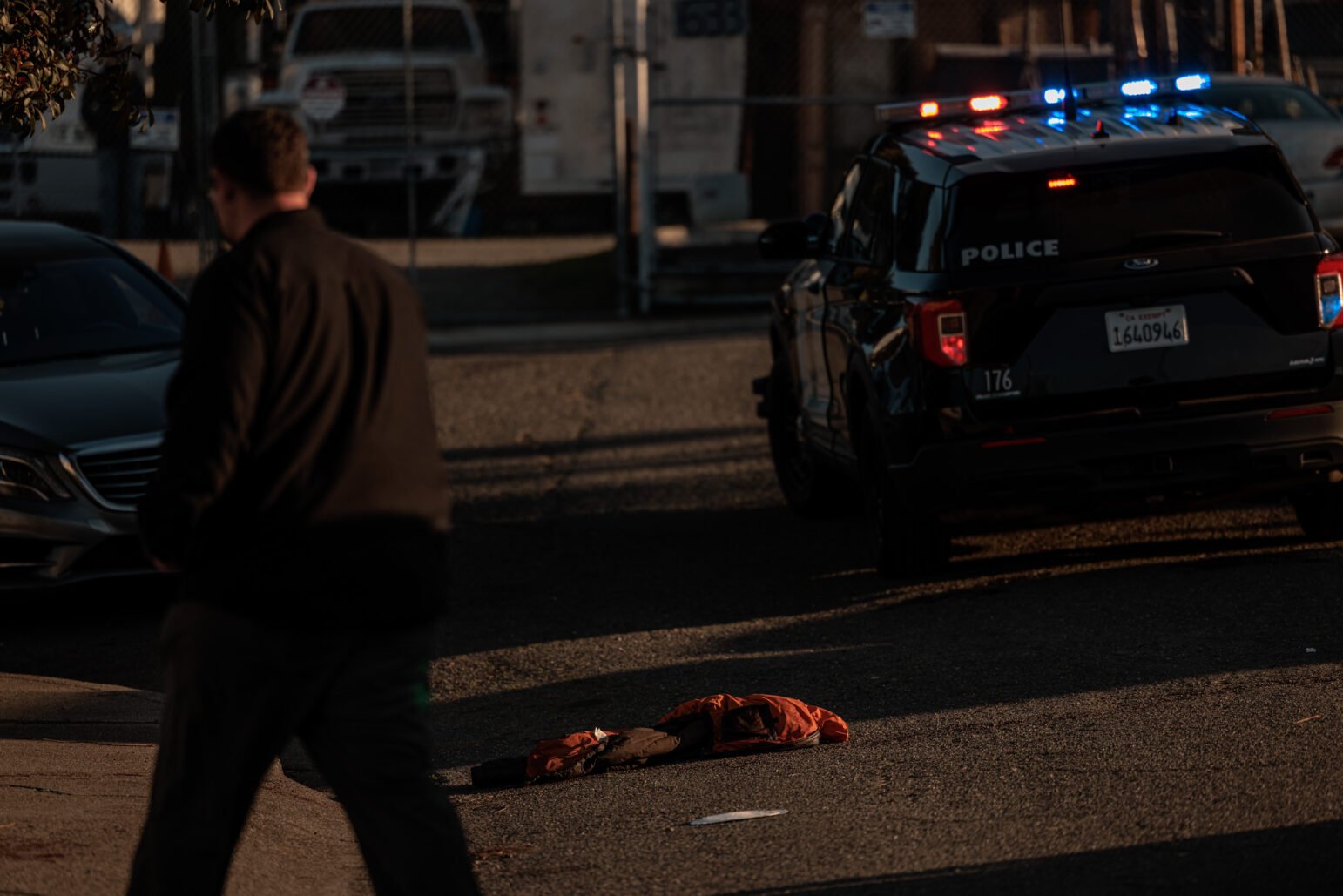 A black police SUV with its emergency lights activated is parked on a sunlit street. In the foreground, a man in dark clothing is out of focus as he walks past the camera. On the pavement between them lies an orange jacket. A chain-link fence and large vehicles are visible in the background.