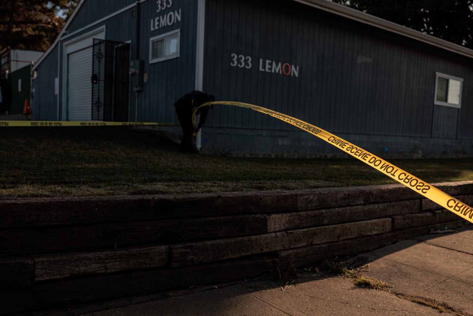 A low‐lit scene outside a gray building bearing the address “333 LEMON.” Yellow crime‐scene tape stretches diagonally across the frame, with a police officer or investigator bent over near the building’s foundation. A grassy lawn slopes up from a wooden retaining wall in the foreground.