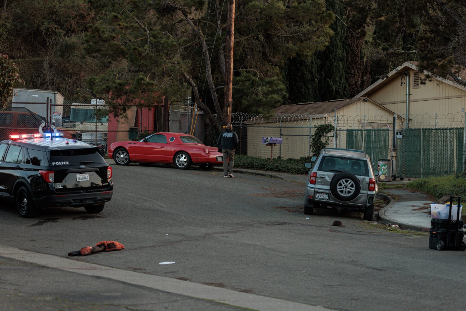 A wide shot of a quiet residential street. In the foreground, a black police SUV with emergency lights activated is stopped near a discarded jacket or vest on the pavement. Further up the street, a red two‐door car is parked, and a police officer—wearing a black jacket labeled “POLICE”—stands near a utility pole. To the right, a silver SUV is parked along the curb. Behind a chain‐link fence topped with barbed wire, a beige single‐story structure and additional vehicles are visible. Trees and hillside foliage fill the background.