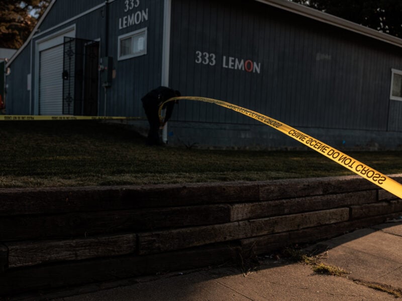 A low‐lit scene outside a gray building bearing the address “333 LEMON.” Yellow crime‐scene tape stretches diagonally across the frame, with a police officer or investigator bent over near the building’s foundation. A grassy lawn slopes up from a wooden retaining wall in the foreground.