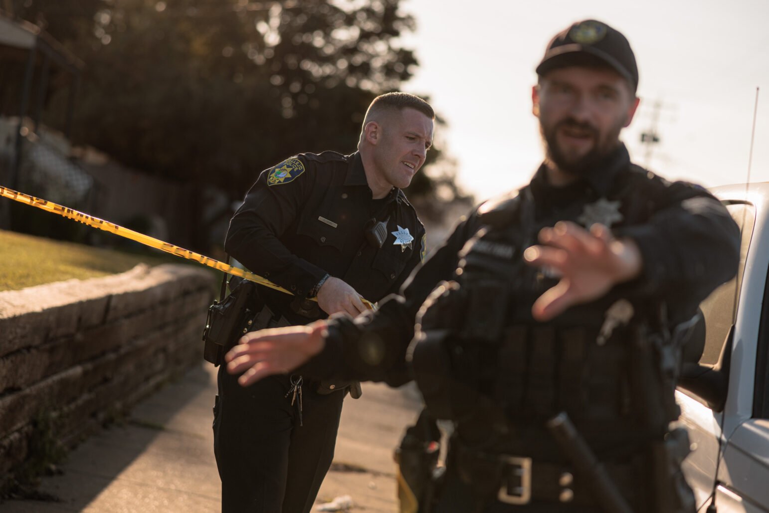 Two uniformed officers in black stand at a cordoned-off area in late-afternoon light. One officer pulls a strip of bright yellow police tape, while the other, out of focus in the foreground, extends a hand toward the camera.