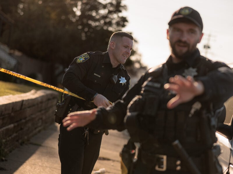 Two uniformed officers in black stand at a cordoned-off area in late-afternoon light. One officer pulls a strip of bright yellow police tape, while the other, out of focus in the foreground, extends a hand toward the camera.