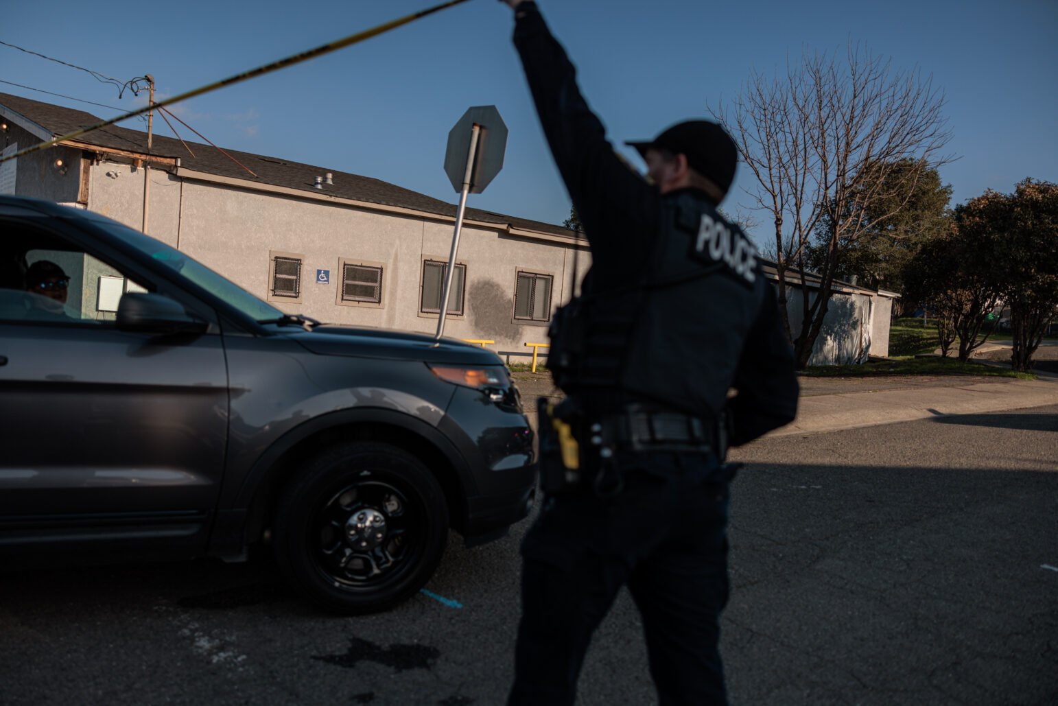 A uniformed police officer, seen from behind, lifts a strip of yellow crime‐scene tape overhead as a dark gray SUV passes underneath. The background shows a one‐story light‐colored building, a wheelchair‐accessible sign, and leafless trees against a bright blue sky.