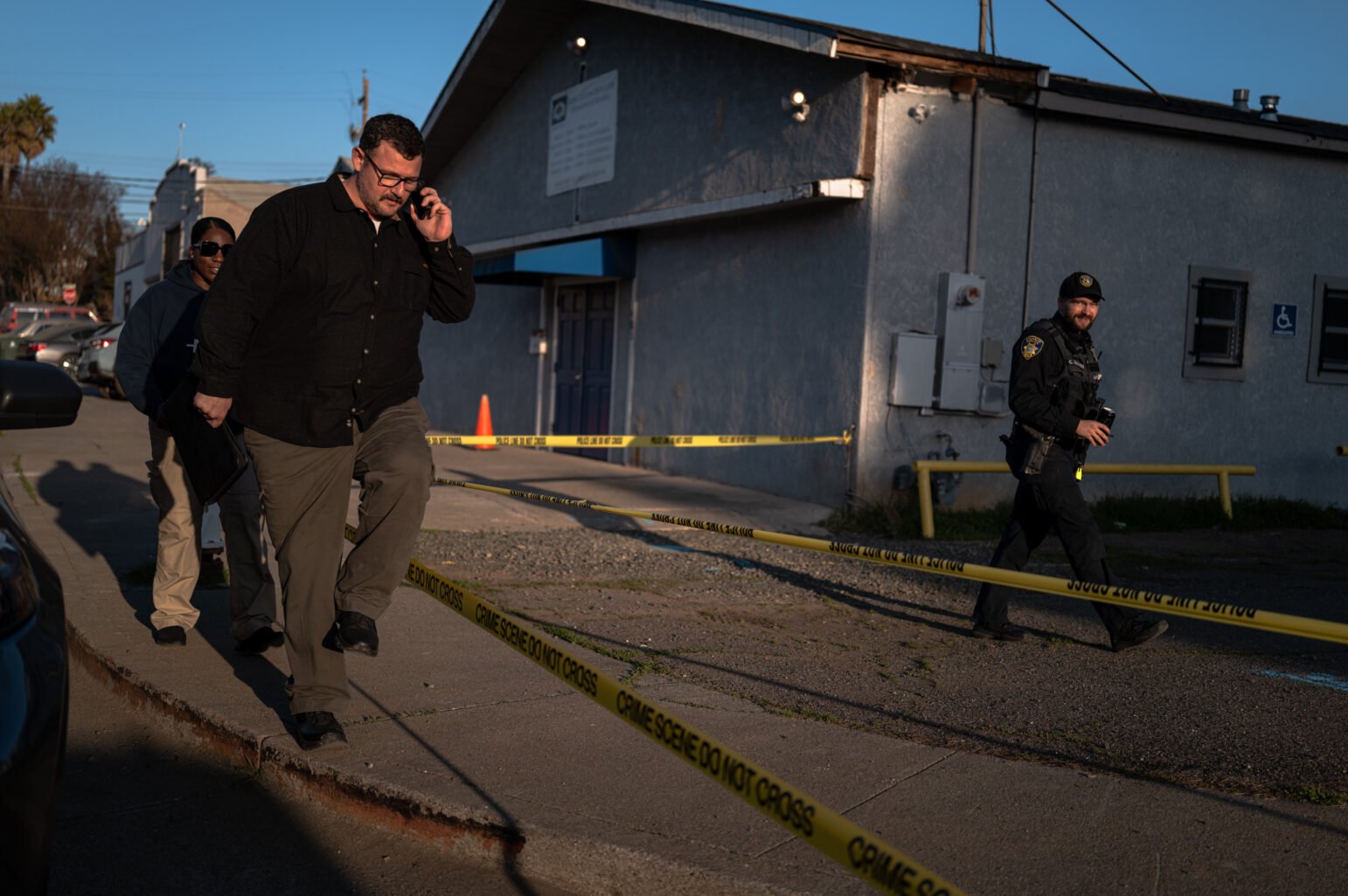 A daytime scene outside a grey building cordoned off by yellow “Crime Scene Do Not Cross” tape. In the foreground, a man wearing a black button-down shirt and tan pants walks over the curb while talking on a phone. Behind him, another individual in dark attire follows closely. At the right edge, a uniformed police officer, wearing a cap and carrying equipment, steps over the tape. The sun casts long shadows on the sidewalk.