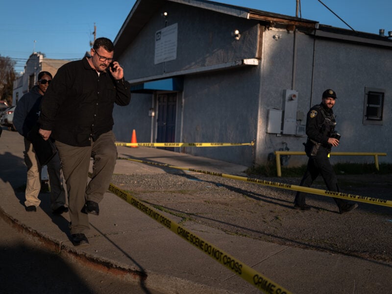 A daytime scene outside a grey building cordoned off by yellow “Crime Scene Do Not Cross” tape. In the foreground, a man wearing a black button-down shirt and tan pants walks over the curb while talking on a phone. Behind him, another individual in dark attire follows closely. At the right edge, a uniformed police officer, wearing a cap and carrying equipment, steps over the tape. The sun casts long shadows on the sidewalk.