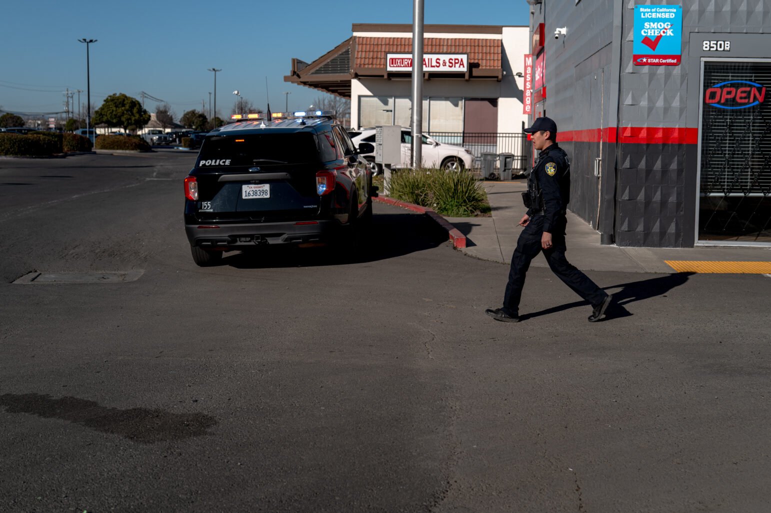 A black police SUV with its emergency lights activated is parked near a nail salon and spa building. A uniformed officer in dark attire strides across the parking lot in the foreground, heading toward the left side of the frame.