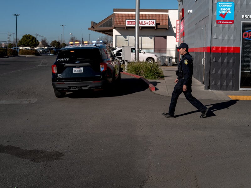 A black police SUV with its emergency lights activated is parked near a nail salon and spa building. A uniformed officer in dark attire strides across the parking lot in the foreground, heading toward the left side of the frame.