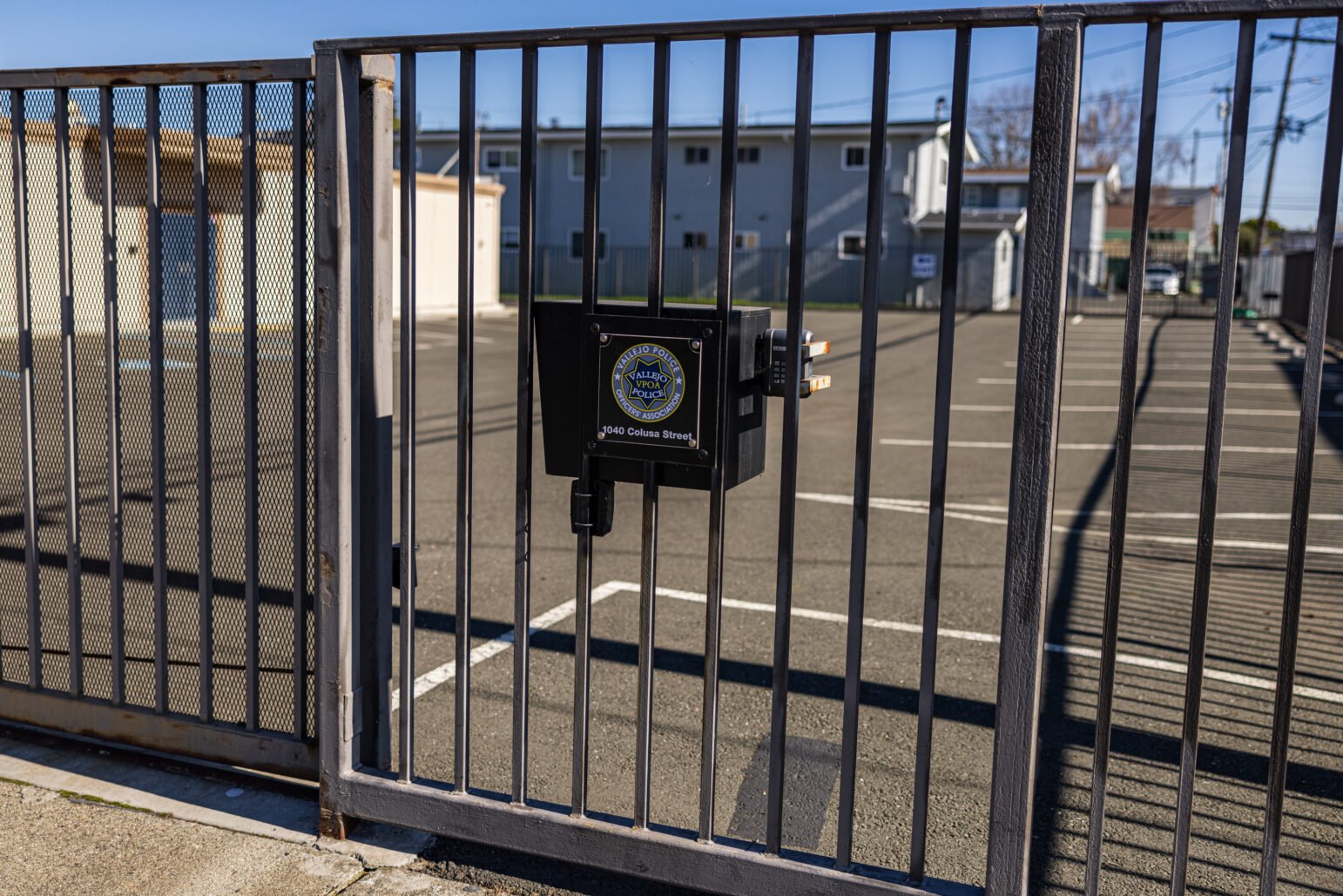 A metal gate enclosing a mostly empty parking lot, with a lockbox on the gate that bears the Vallejo Police Officers’ Association badge and the address “1040 Colusa Street.” In the background, multiple parking stalls and a row of buildings are visible under a clear, blue sky.