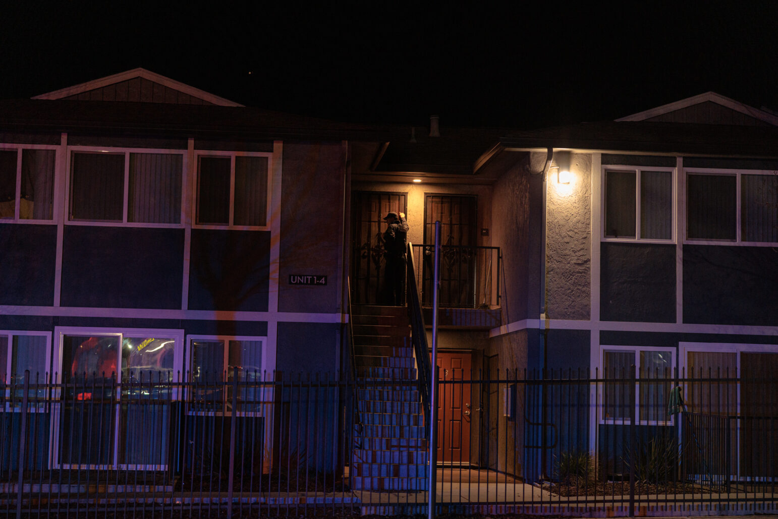 A two-story apartment building at night. An officer in dark uniform stands on the external staircase landing, knocking on or examining a door under a single porch light.