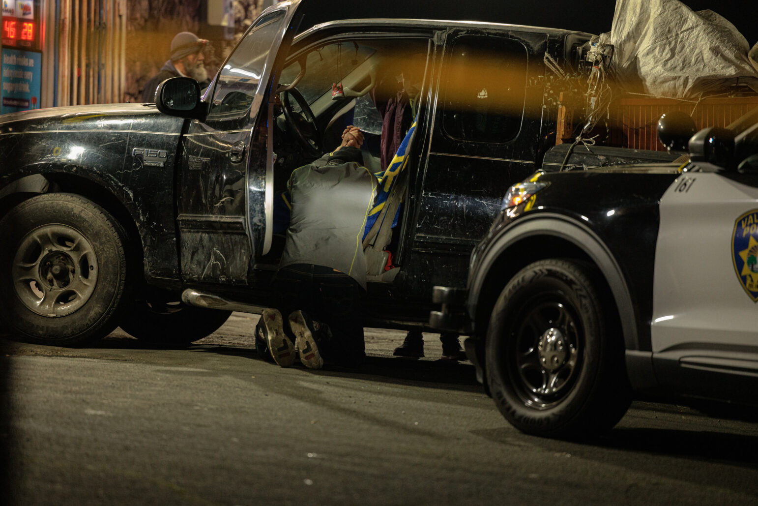 Under bright artificial light, the driver’s door of a black Ford F-150 is open. A person leans forward on their knees inside the cab, while another individual wearing a beanie stands outside. A marked police SUV is parked nearby. The truck’s paint appears worn, and various items are strapped to its bed.